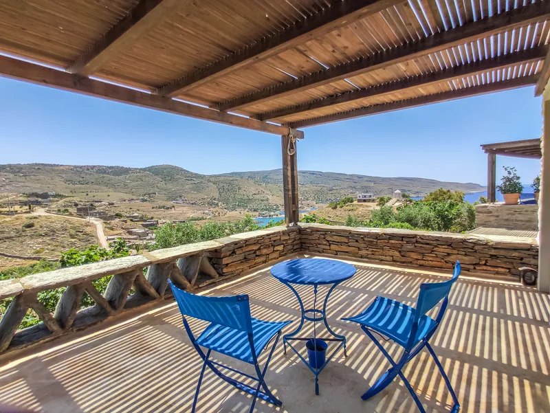 Pergola terrace with blue bistro chairs and the Aegean and Koundouros bay in bright morning light
