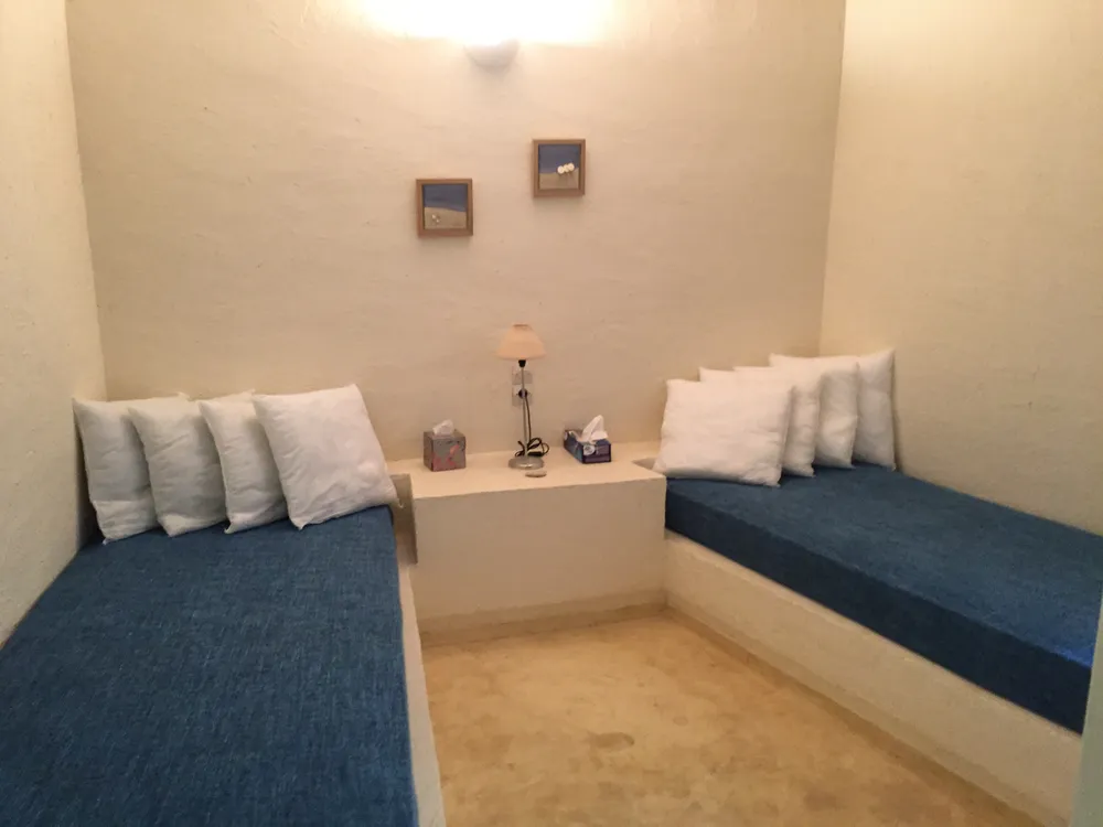 Two blue-cushioned single beds on plaster bases with white pillows, small framed prints above, and a central lamp