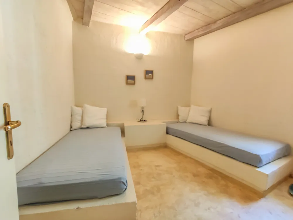 Two single beds on whitewashed plaster platforms with grey mattresses, shared plaster bedside table, and a wall lamp