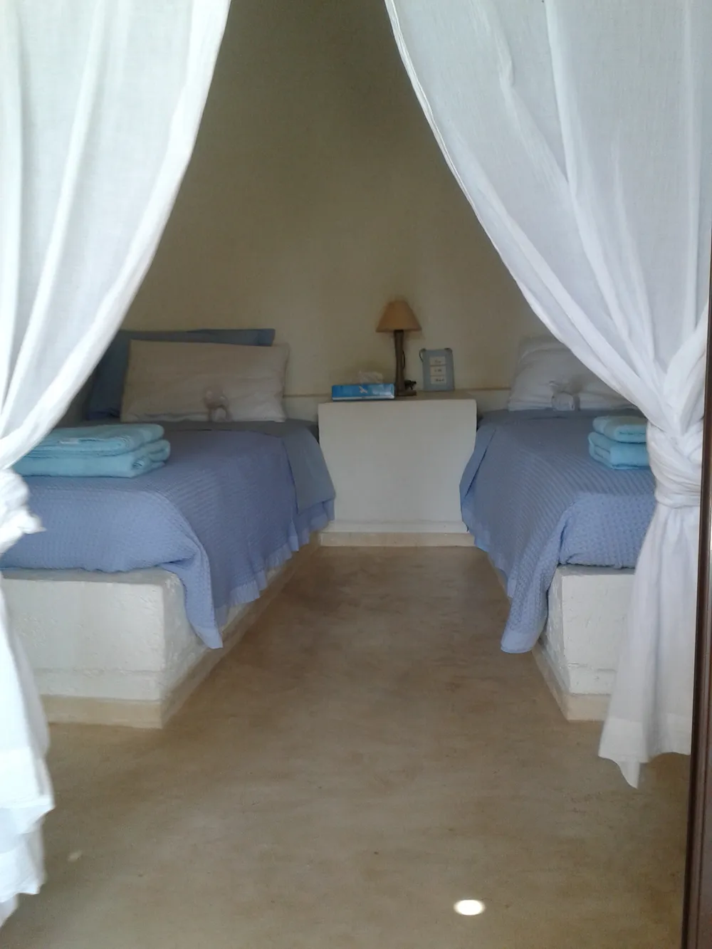 Twin bedroom with two built-in plaster-base beds in blue covers, framed by sheer white curtains at the doorway
