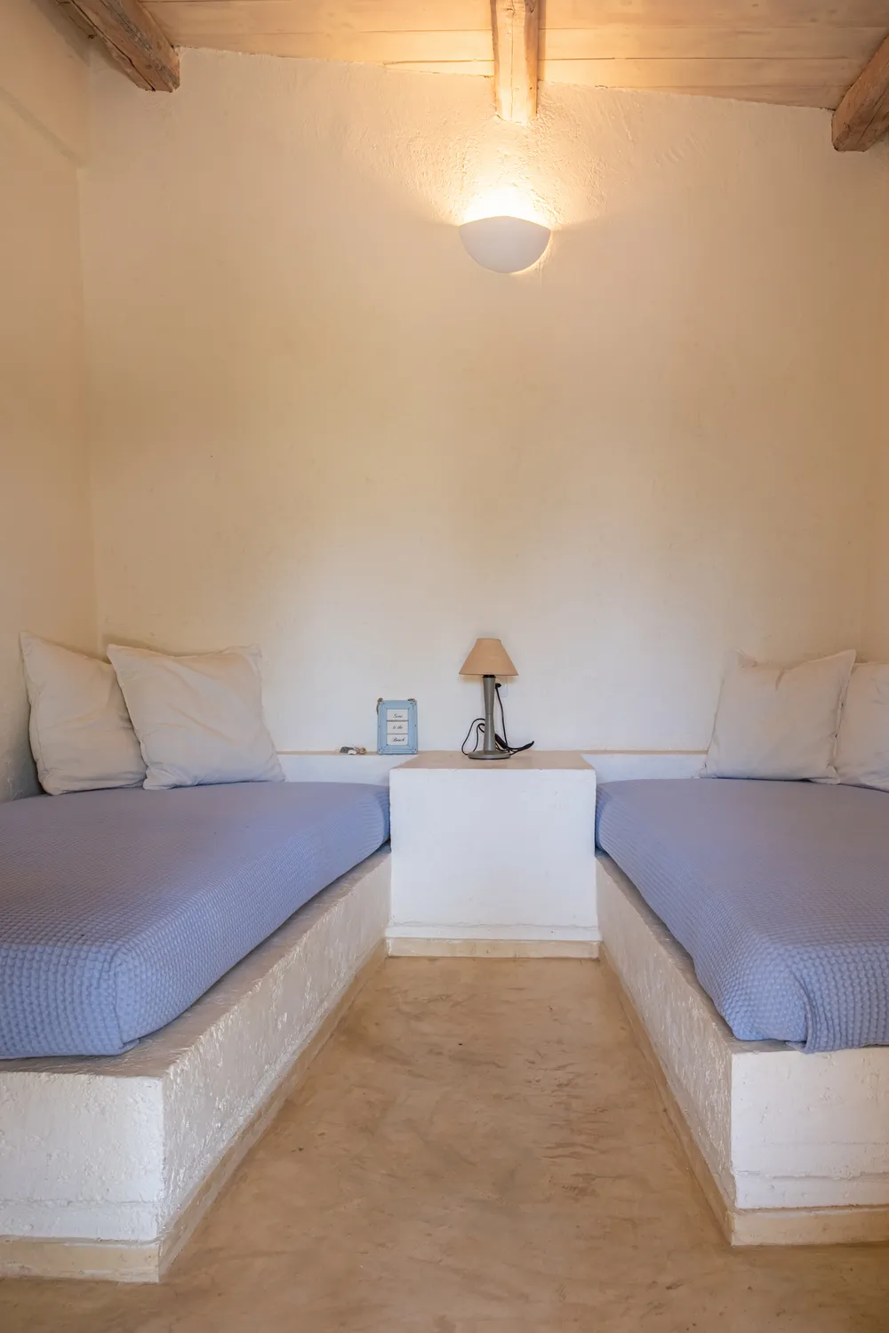 Twin bedroom with two plaster-base beds and blue covers, plaster walls, exposed wood ceiling beams, and a wall sconce