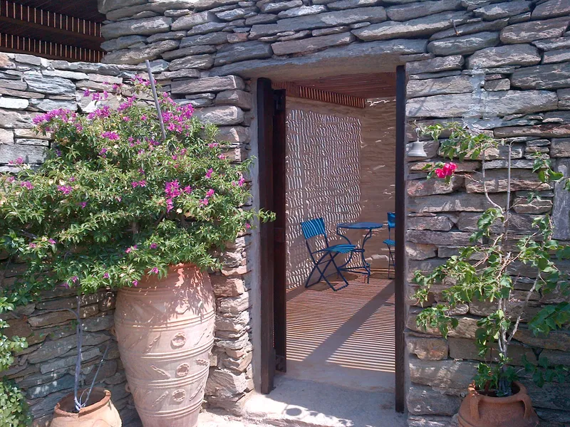 Stone doorway framed by bougainvillea and a tall terracotta urn, opening to a terrace with blue bistro chairs