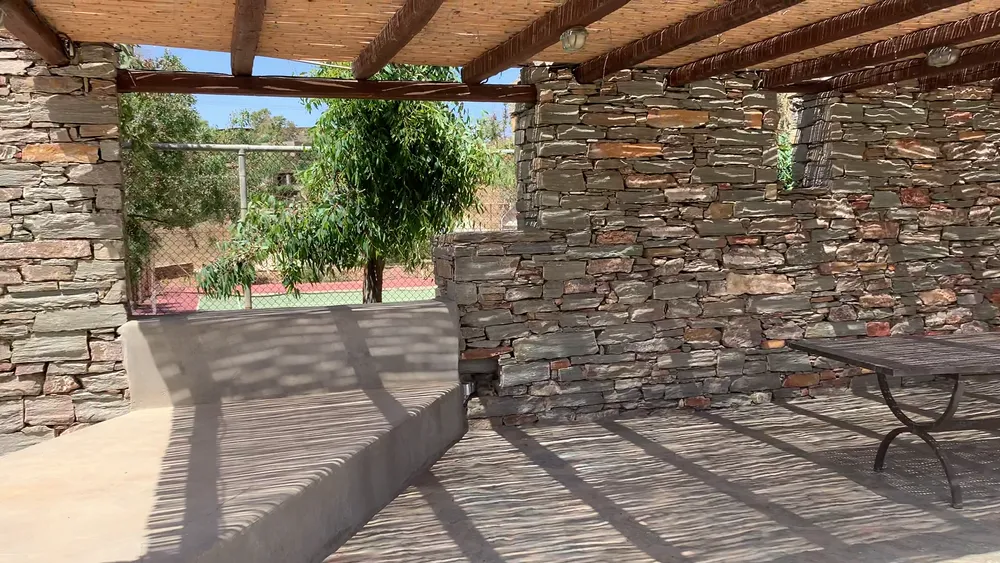 Shaded seating area with dry-stone walls and a reed pergola roof, overlooking the tennis court