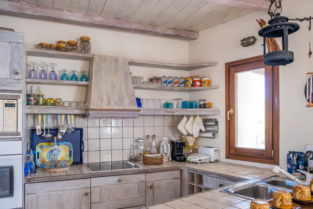 Kitchen cooking corner with open shelves of glassware, tiled splashback, hob, and a window with wooden shutters