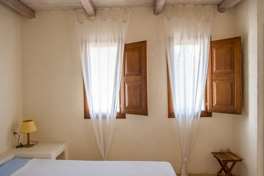 Master bedroom window wall with two sets of wooden shutters, sheer curtains, yellow lamp, and a small wooden stool