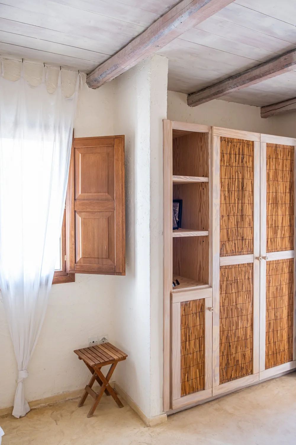 Master bedroom wardrobe with reed-fronted doors and open shelves beside a wooden-shuttered window and sheer curtain