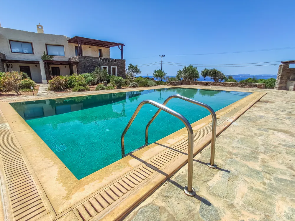 Pool with stainless steel ladder, stone-and-render villa buildings behind, and the sea in the distance